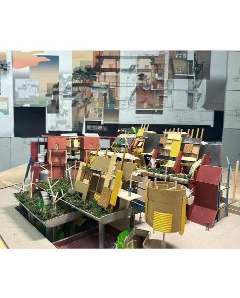 Architectural scale model of colourful stacked structures and plants, in front of a pinned collage wall.