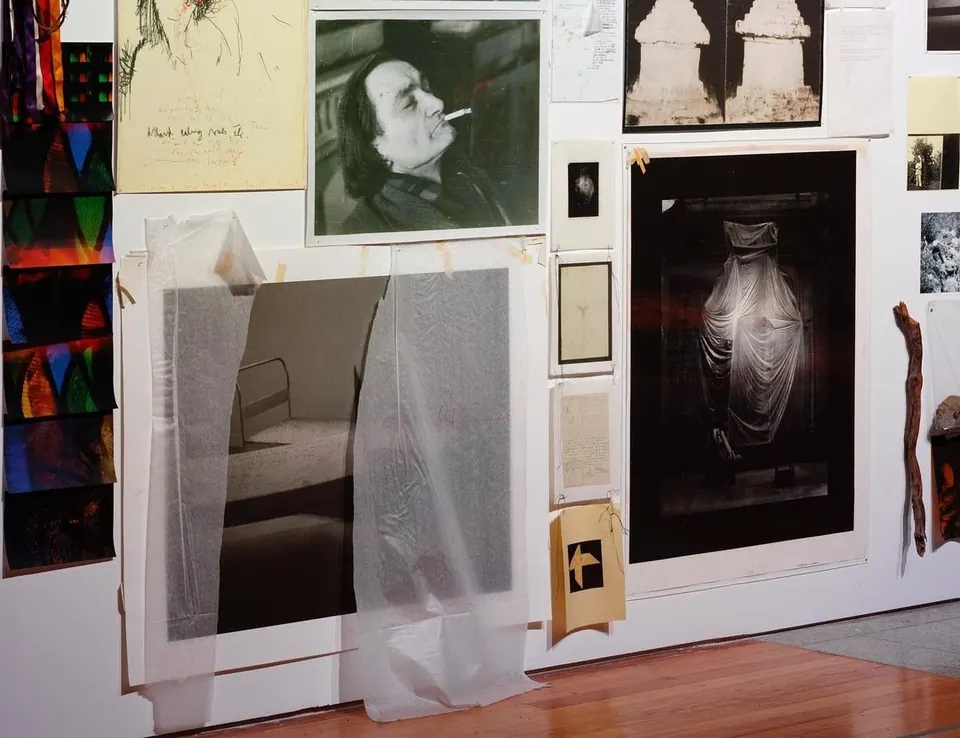 Artist studio wall layered with photographs, sketches, tracing paper and a black-and-white portrait of a smoking man.