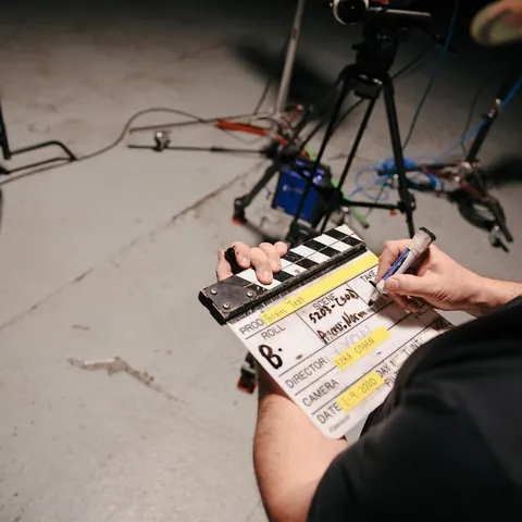 Hands writing scene details on a film clapperboard on set, tripod and cables behind.