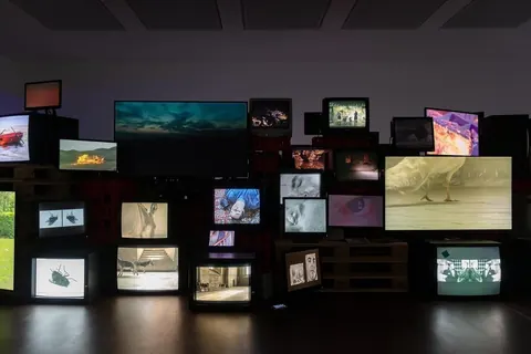 Dark gallery filled with a wall of old CRT TVs and monitors playing different video clips.