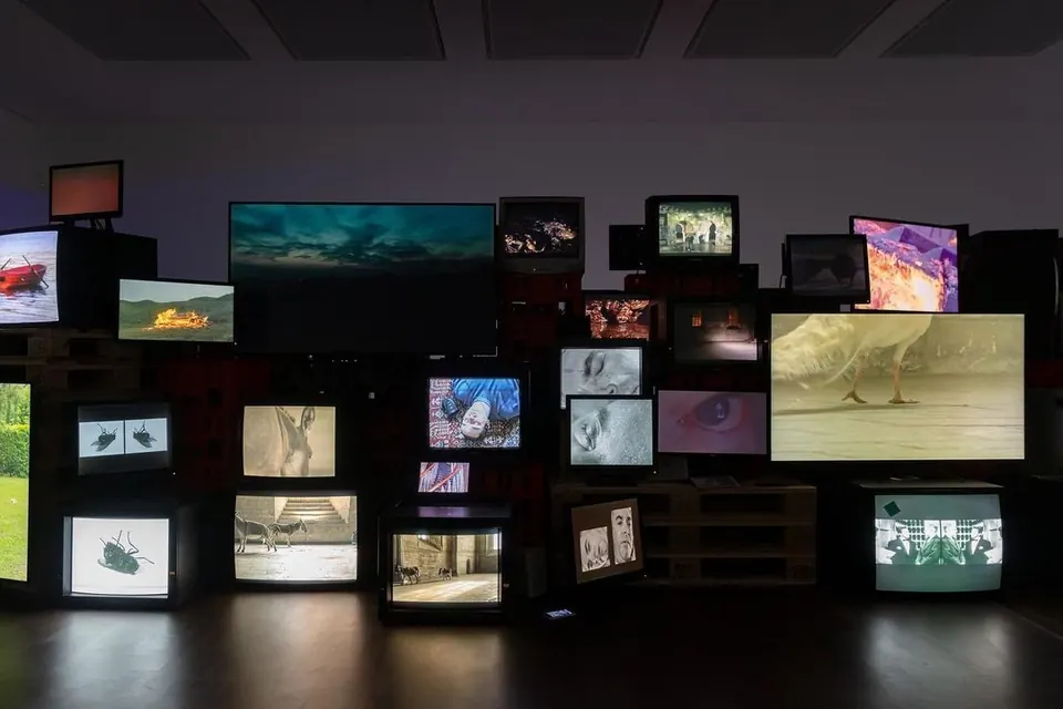 Dark gallery filled with a wall of old CRT TVs and monitors playing different video clips.