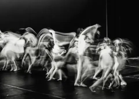 Black-and-white long-exposure photograph of dancers moving across a stage, bodies blurred into ghostly trails.