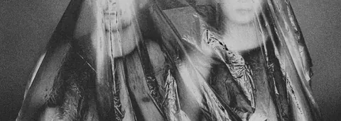 Grainy black-and-white of two faces behind crumpled plastic.