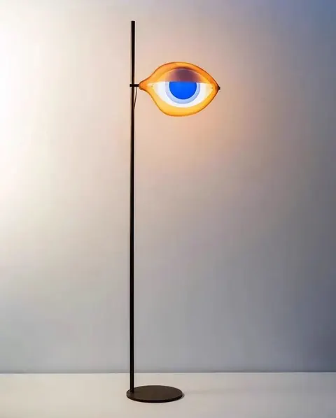 Floor lamp whose glowing shade is shaped like an orange and blue eye, against a pale wall.