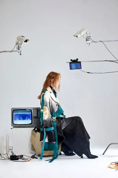 Fashion shoot: model in an oversized teal coat beside a CRT monitor showing an eye, CCTV cameras overhead, white studio.