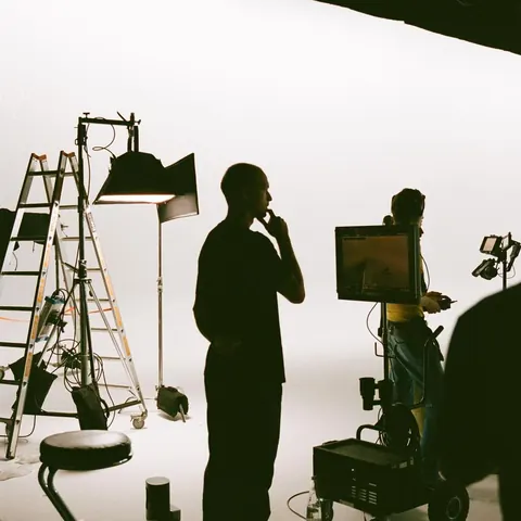 Behind-the-scenes on a film set: silhouetted crew, lights, ladder and a monitor cart in a white cyclorama studio.