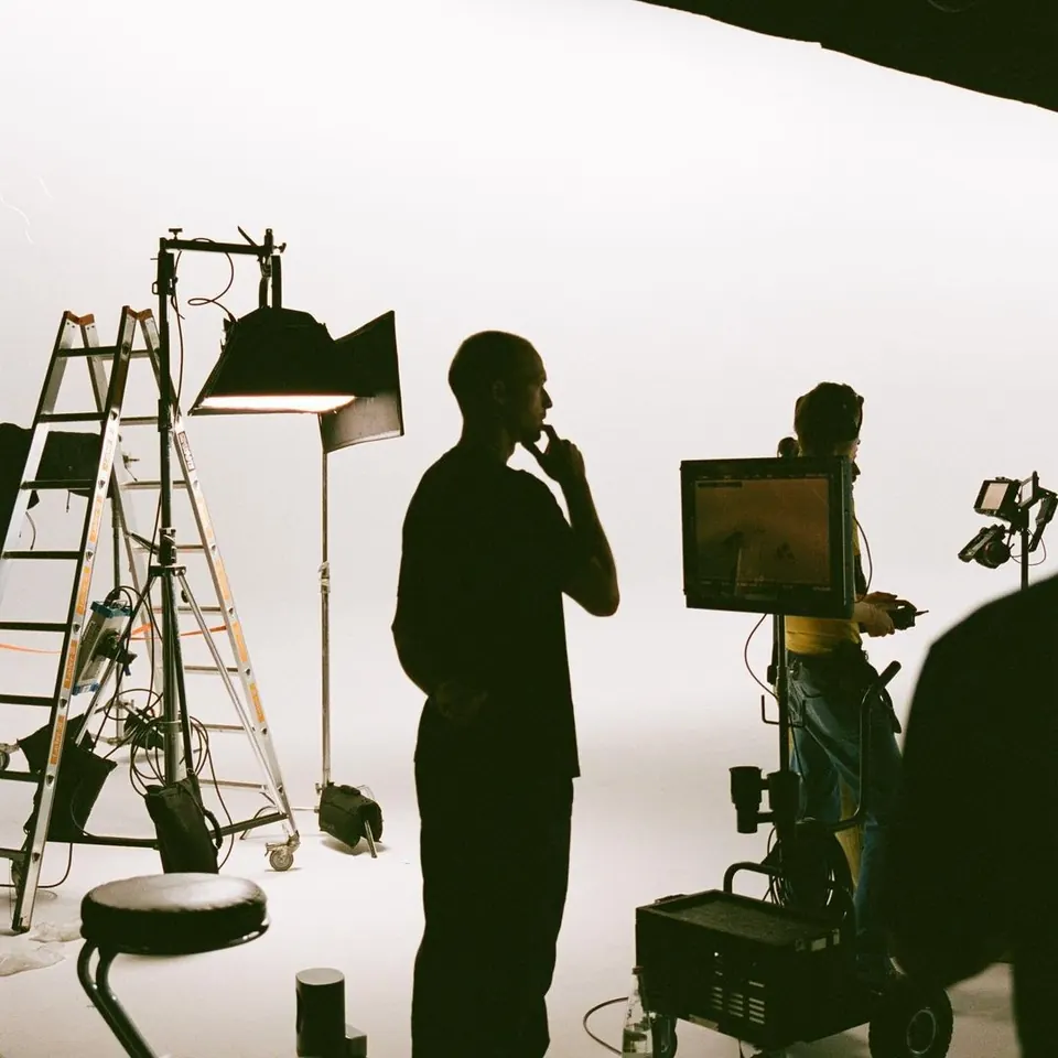 Behind-the-scenes on a film set: silhouetted crew, lights, ladder and a monitor cart in a white cyclorama studio.