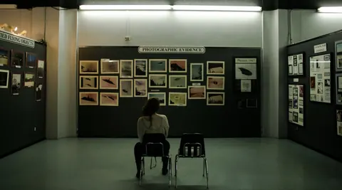 A lone viewer seated in a gallery hung with small framed photographs under a "Photographic Evidence" sign.