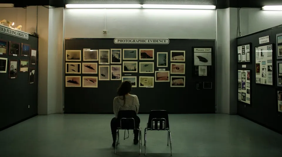 A lone viewer seated in a gallery hung with small framed photographs under a "Photographic Evidence" sign.