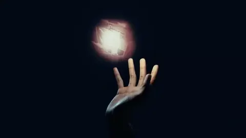 Hand reaching up toward a glowing cube of light in the dark.