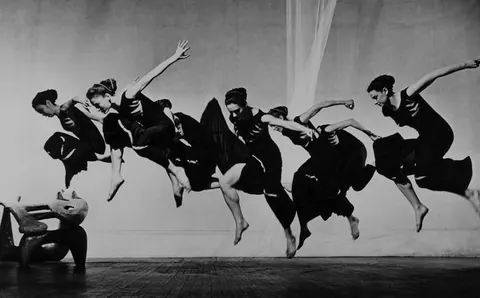 Black-and-white photo of a line of dancers in dark dresses leaping in unison.