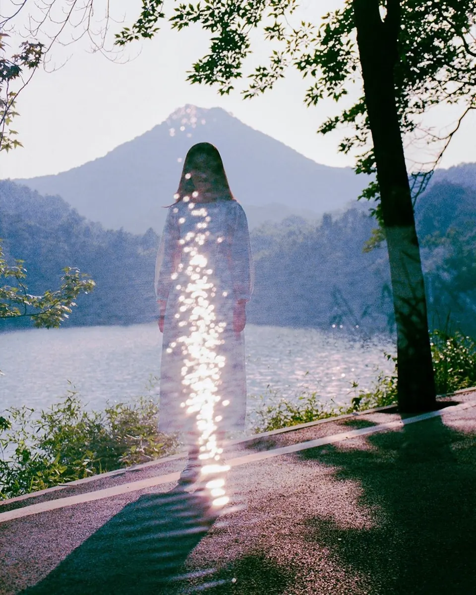 Film photograph of a figure in white dissolved by sparkling light, by a lake with a mountain behind.