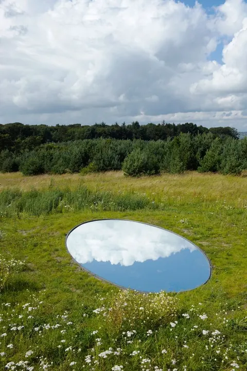 Land-art piece: an oval mirror lying in a meadow, reflecting sky and clouds.