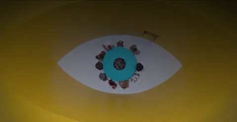 Overhead shot of people seated around a teal table inside a giant painted eye shape on a yellow floor.