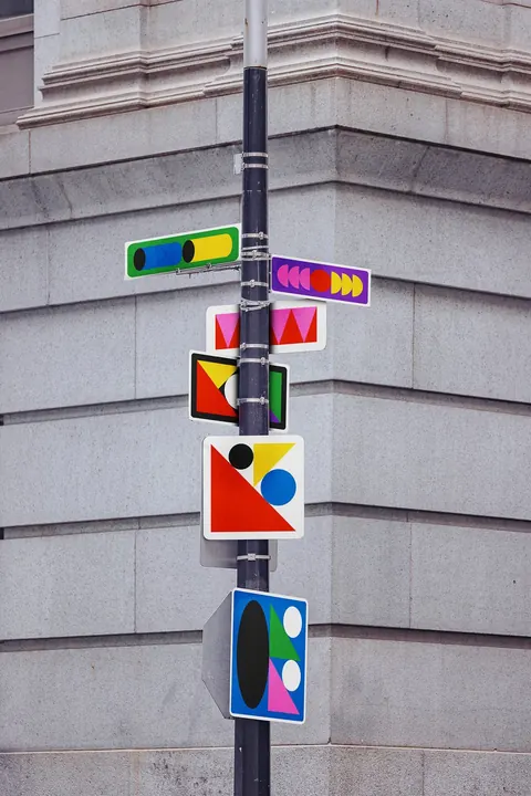 Street pole on a stone building fitted with abstract geometric painted signs in red, yellow, blue and green.