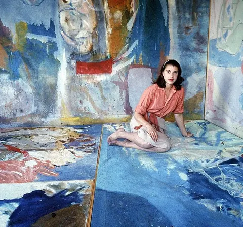 Portrait of a woman seated on the floor of a room whose walls and floor are covered in expressive blue and red paint.