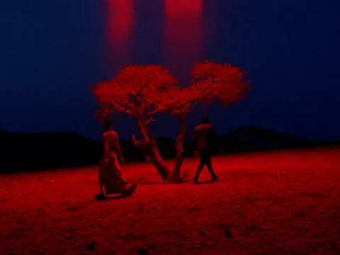 Cinematic still: two figures walking past a red-lit tree on red earth under a deep blue night sky.