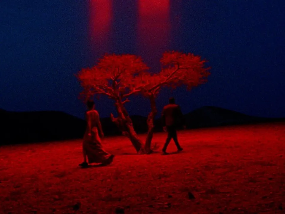 Cinematic still: two figures walking past a red-lit tree on red earth under a deep blue night sky.