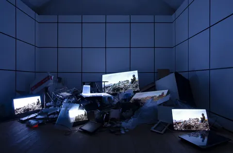 Dark blue room installation: a heap of old computer monitors and screens glowing with landscape footage.