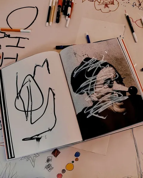 Open book with scribbled drawings over a photo, markers scattered on a desk.