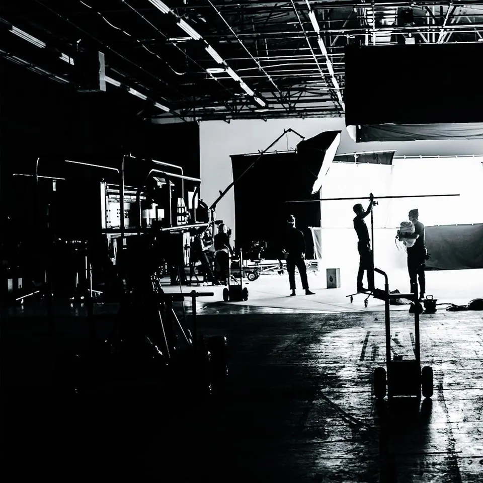 Black-and-white photo of a large soundstage: crew silhouettes against a bright white backdrop, lighting rigs overhead.