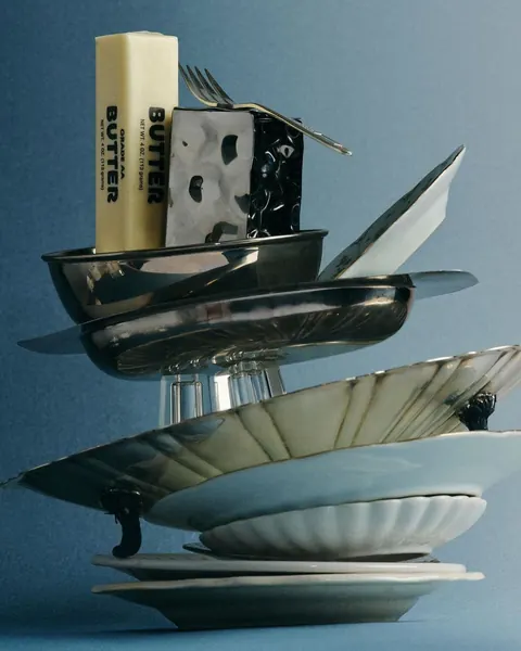 Still life of stacked silver plates, dishes, a stick of butter and cheese against blue.