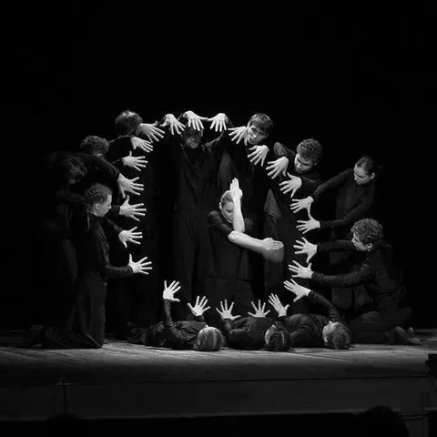 Black-and-white theatre photograph: performers in black form a circle of outstretched hands around a central figure.