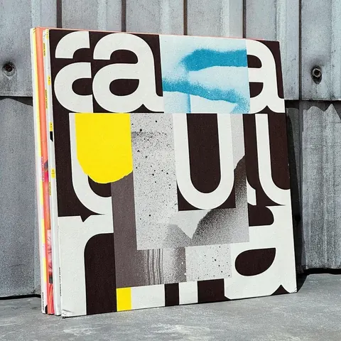 Large typographic print of cropped letterforms "a", "u", "r" with yellow, blue and grey textures, leaning against a corrugated wall.